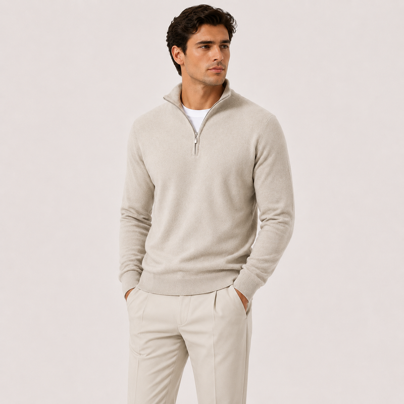 A man wearing a light beige quarter-zip knit sweater over a white undershirt and matching beige trousers, standing against a plain light beige studio background.