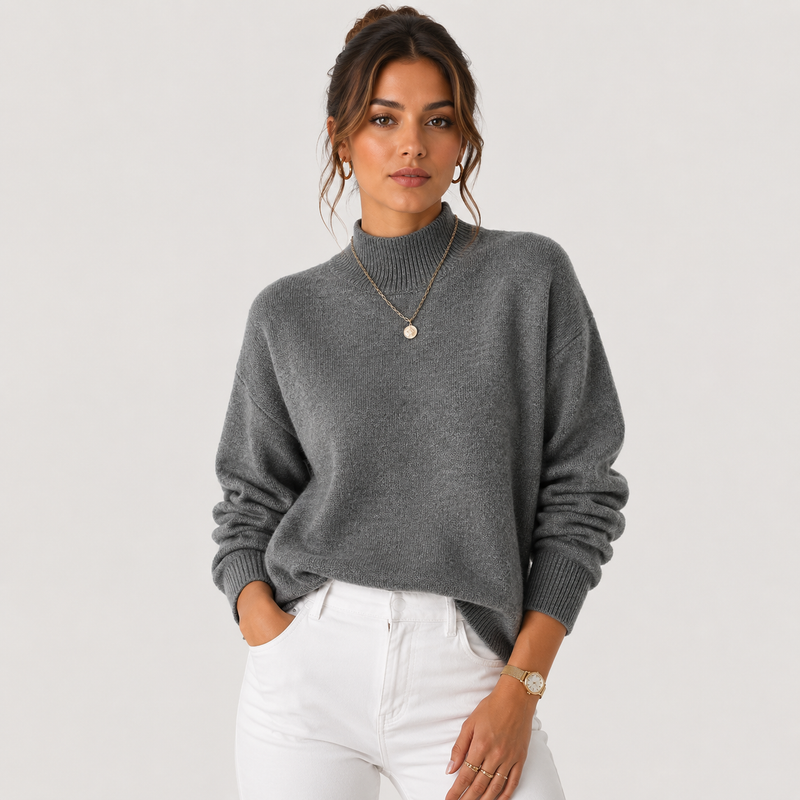 A woman wearing a gray ribbed mock neck sweater and white pants, standing against a light gray studio background.