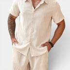 Man wearing a beige shirt and pants with tattoos on arms against a white background