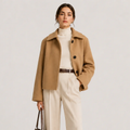 A woman models a camel-colored wool-blend coat over a cream turtleneck and cream wide-leg trousers, accessorized with a brown belt and holding a dark brown handbag, set against a plain light gray studio background.