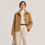 A woman models a camel-colored wool-blend coat over a cream turtleneck and cream wide-leg trousers, accessorized with a brown belt and holding a dark brown handbag, set against a plain light gray studio background.