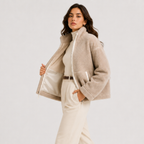 A woman models a beige sherpa fleece zip-up jacket with a high collar, worn open over a cream mock-neck top and cream trousers, against a plain light gray studio background.