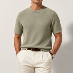 A man wearing a short-sleeve textured knit crewneck sweater in sage green, paired with off-white trousers and a brown leather belt, photographed against a neutral light gray studio background.