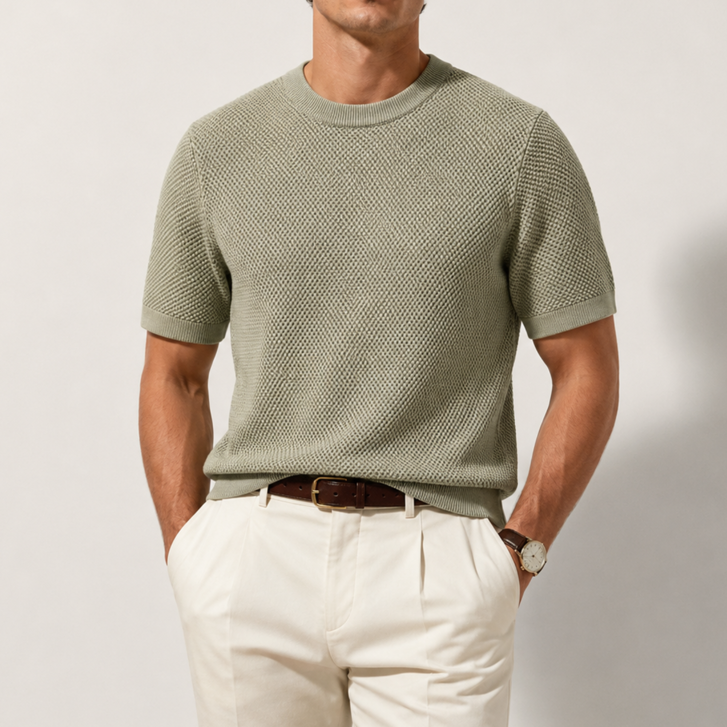 A man wearing a short-sleeve textured knit crewneck sweater in sage green, paired with off-white trousers and a brown leather belt, photographed against a neutral light gray studio background.