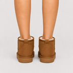 A pair of brown suede ankle boots with white shearling lining, shown from the back on a model's feet against a light gray background.
