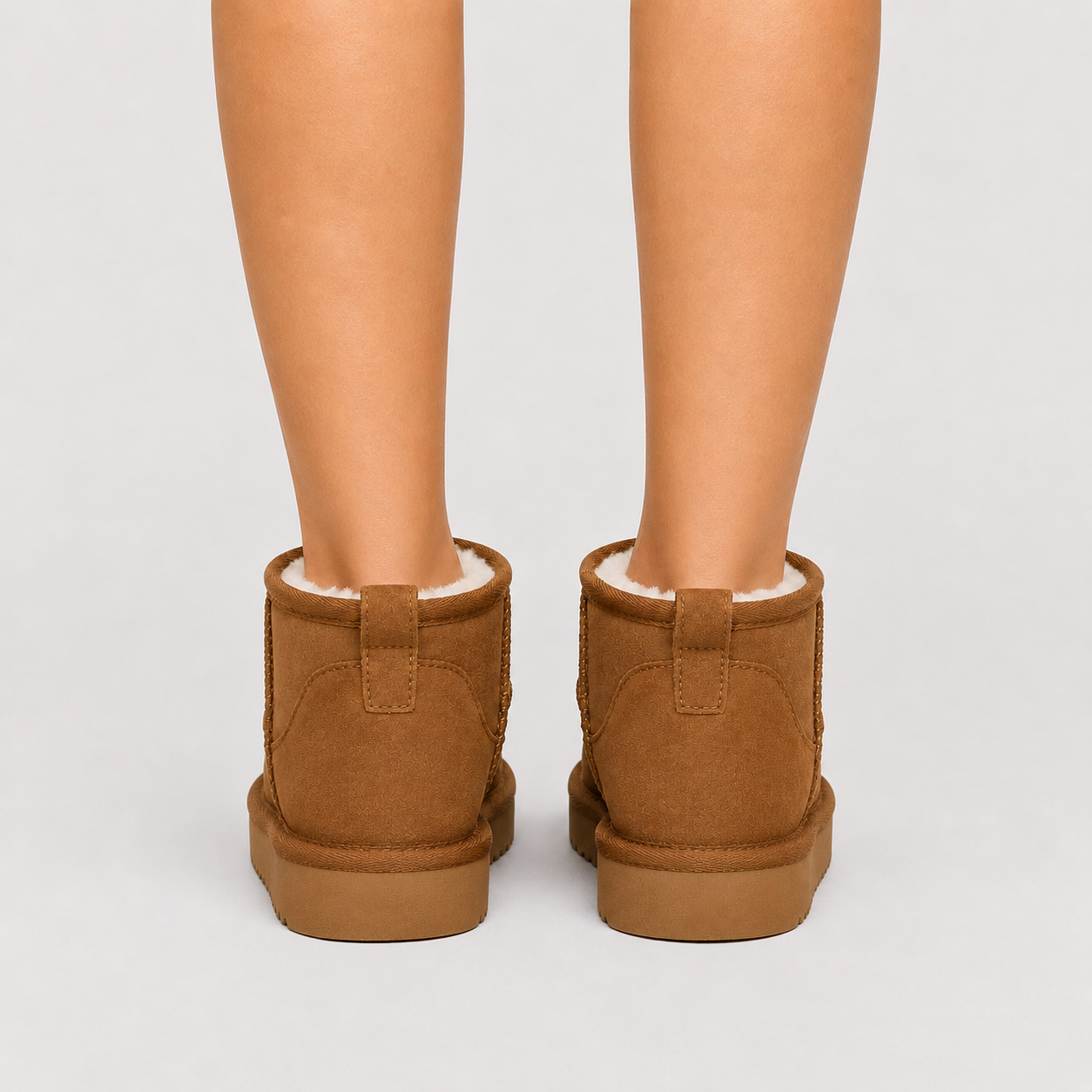 A pair of brown suede ankle boots with white shearling lining, shown from the back on a model's feet against a light gray background.