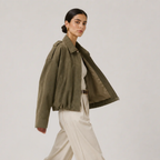 A woman models an olive green cropped utility jacket with voluminous sleeves, worn open over a white top and cream wide-leg trousers, set against a plain light gray studio background.