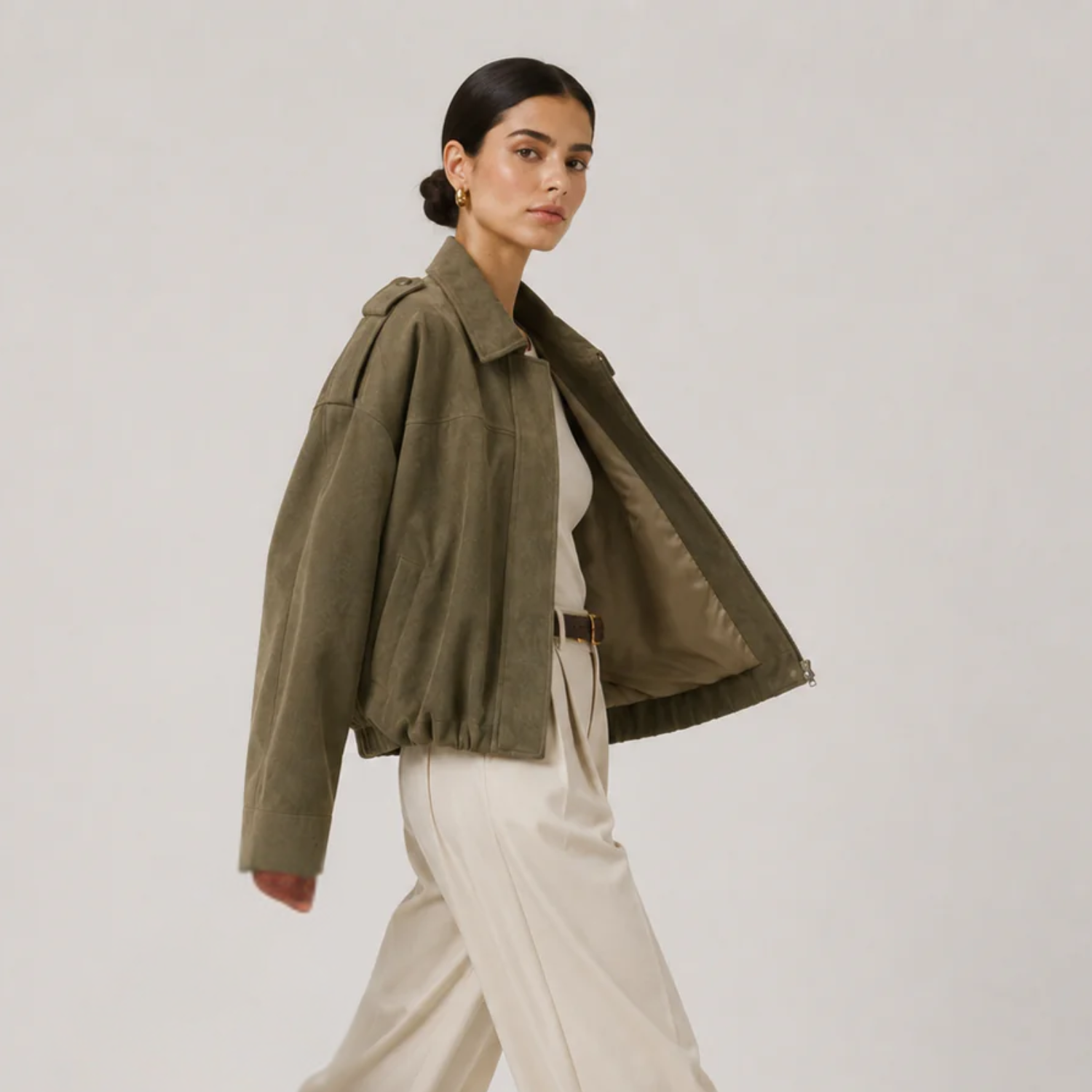 A woman models an olive green cropped utility jacket with voluminous sleeves, worn open over a white top and cream wide-leg trousers, set against a plain light gray studio background.