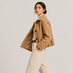 A woman modeling a cropped camel-colored coat over a cream turtleneck and cream trousers, photographed against a plain light gray studio background.