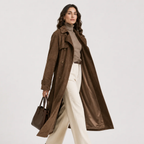A woman models a long brown suede trench coat over a taupe turtleneck and cream wide-leg trousers, holding a matching brown leather handbag against a light gray studio background.