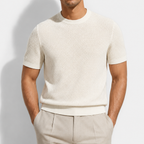 A man wearing a short-sleeve knit crewneck sweater in cream, paired with light beige trousers, shown from the waist up against a light gray background.