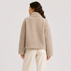 A woman shown from the back wearing a beige sherpa fleece cropped jacket and cream-colored pants, photographed against a clean light gray studio background.