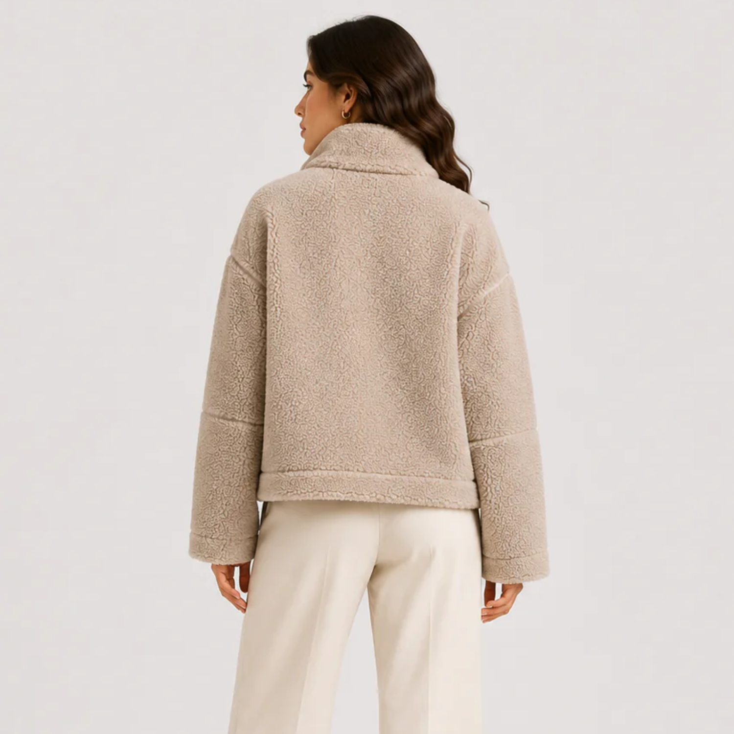 A woman shown from the back wearing a beige sherpa fleece cropped jacket and cream-colored pants, photographed against a clean light gray studio background.