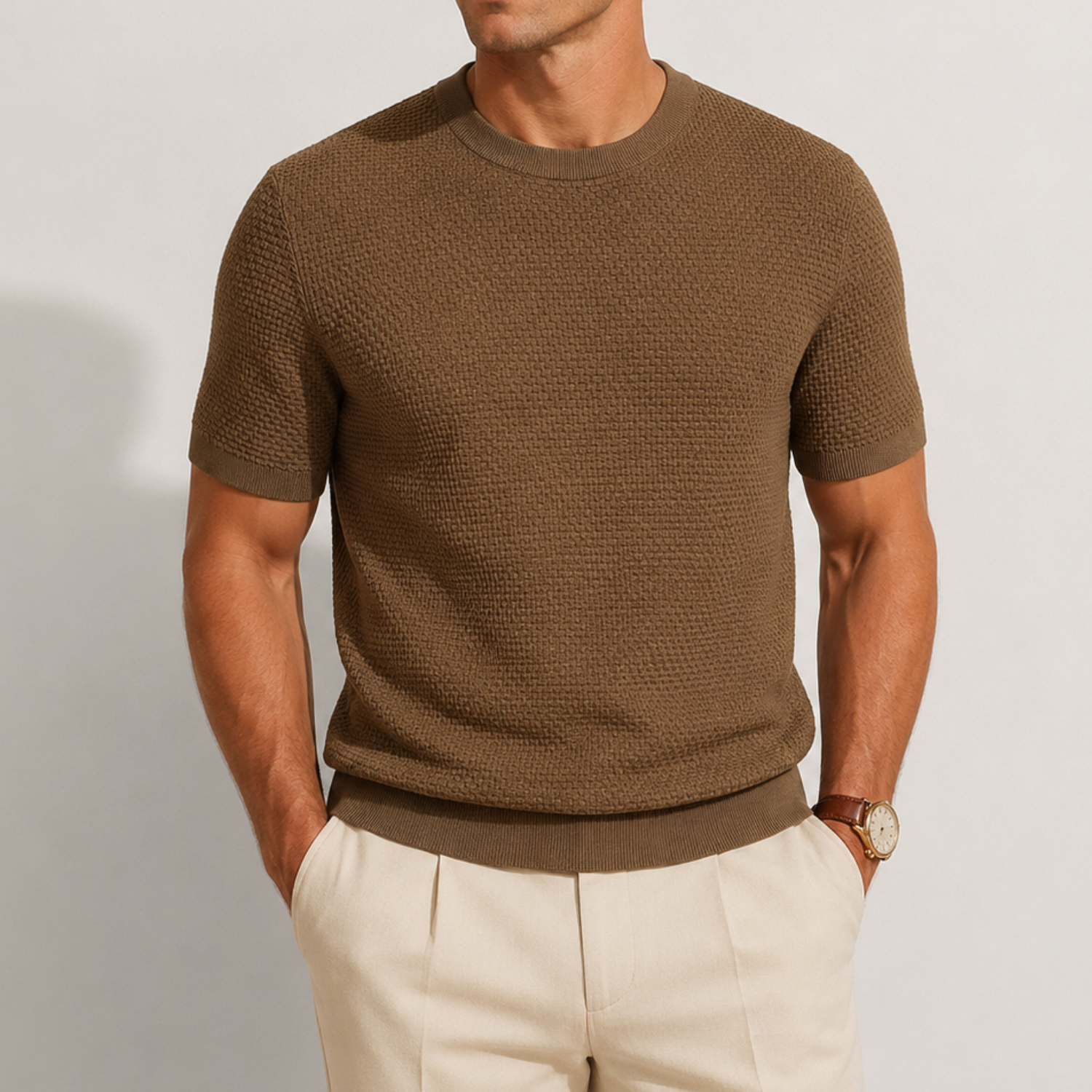 A man wearing a textured short-sleeve crewneck knit sweater in brown, paired with light beige trousers, shown from the chin down against a plain light gray background.