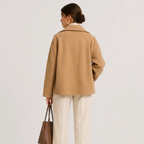 A woman shown from the back wearing a camel-colored oversized coat, cream trousers, and holding a brown handbag against a plain light gray studio background.