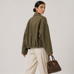A woman wearing an oversized olive green jacket, cream wide-leg trousers, and holding a brown leather handbag, photographed against a plain light gray studio background.