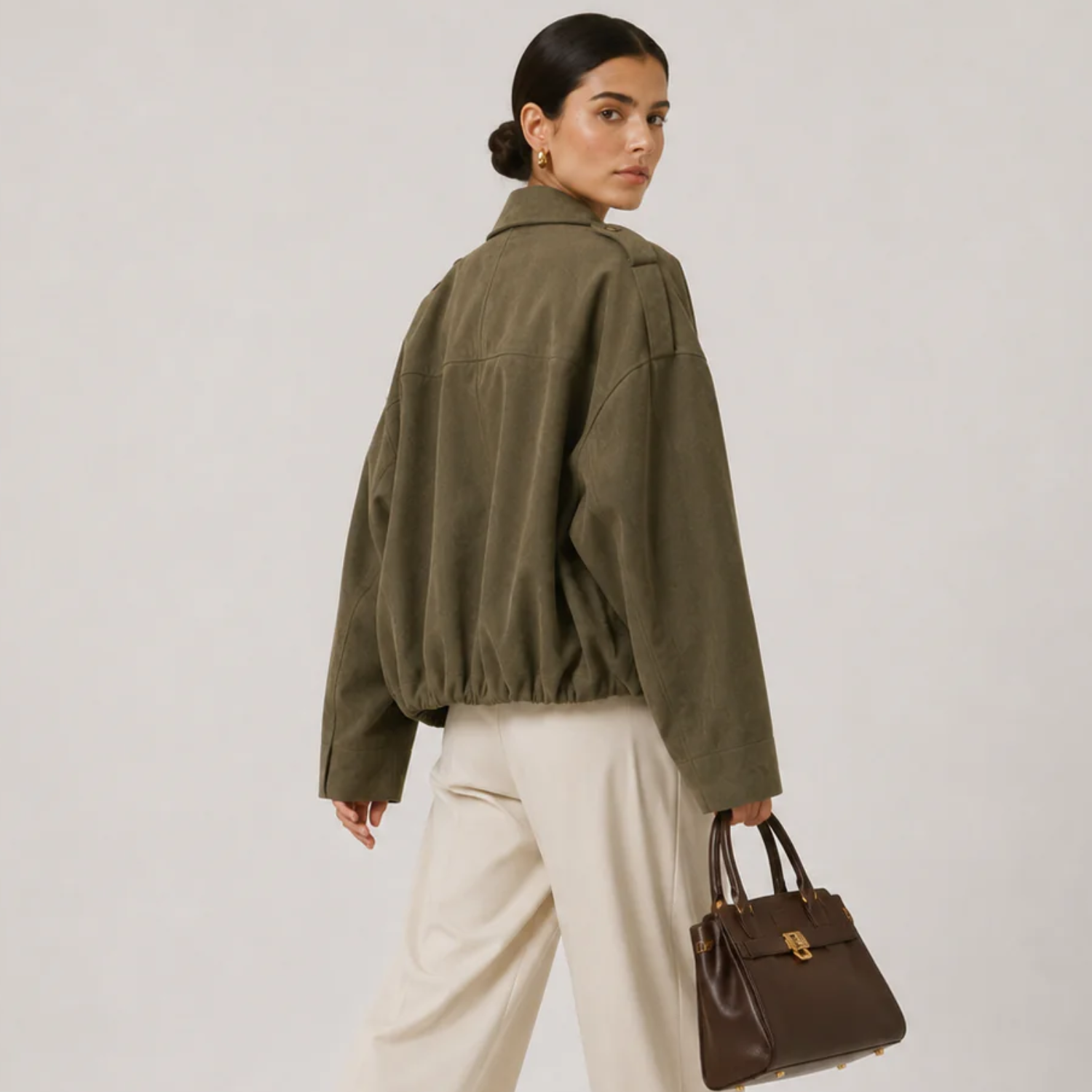 A woman wearing an oversized olive green jacket, cream wide-leg trousers, and holding a brown leather handbag, photographed against a plain light gray studio background.