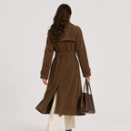 A woman viewed from behind wearing a brown suede trench coat with a matching belt, paired with cream pants and holding a dark brown leather handbag against a light gray studio background.