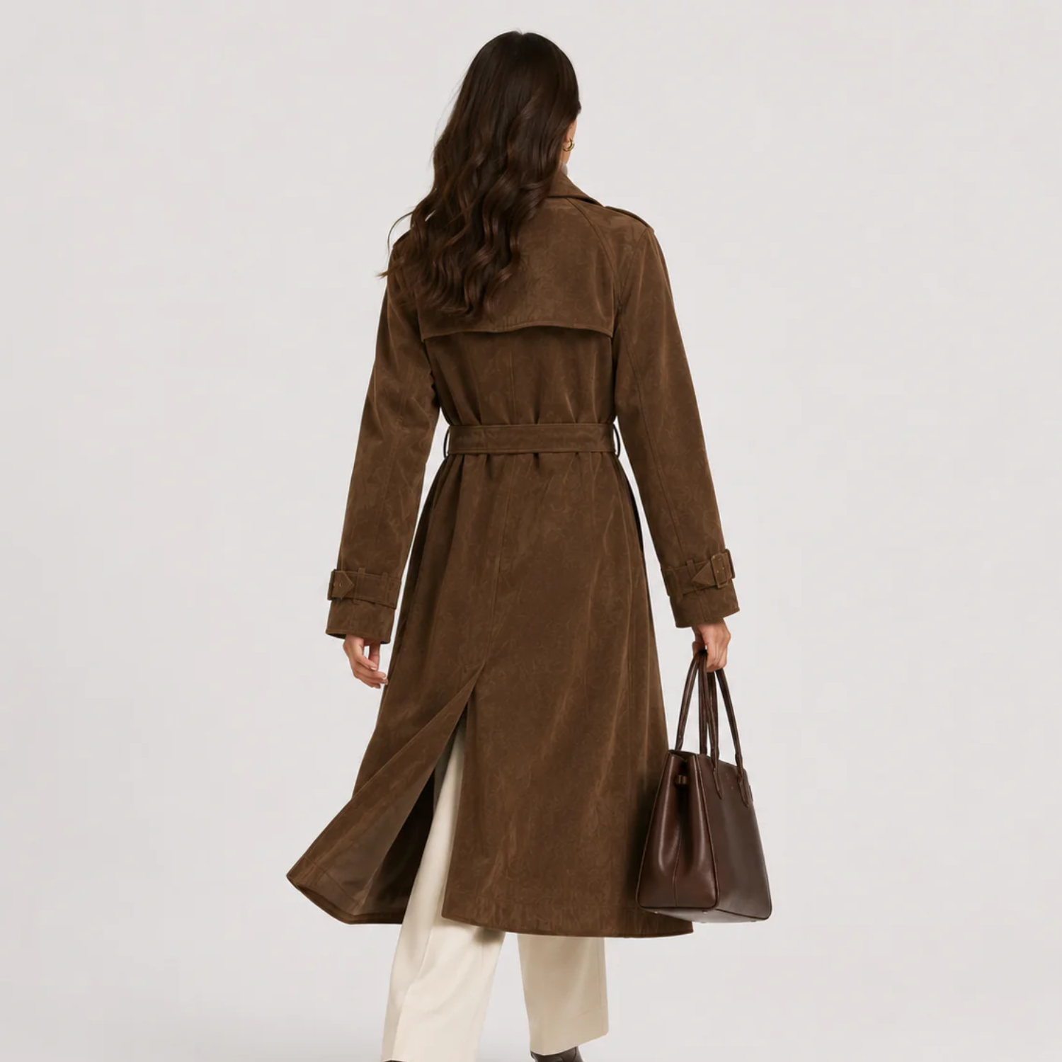 A woman viewed from behind wearing a brown suede trench coat with a matching belt, paired with cream pants and holding a dark brown leather handbag against a light gray studio background.