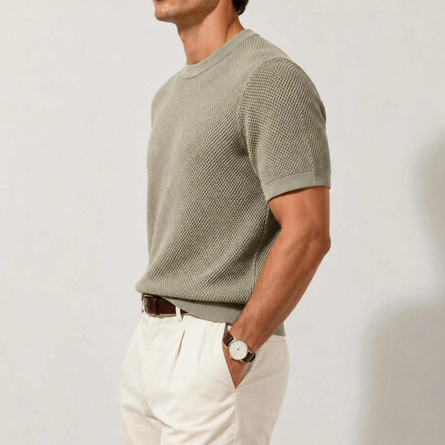 A man wearing a short-sleeve textured knit crewneck sweater in sage green, paired with off-white trousers and a brown leather belt, shown from the side against a plain light gray background.