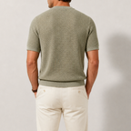 A man shown from the back wearing a textured sage green short-sleeve knit shirt and off-white trousers, standing against a neutral light gray studio background.
