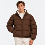 A man wearing a brown puffer jacket with a high collar over a white ribbed turtleneck sweater and off-white trousers, photographed against a clean light gray studio background.