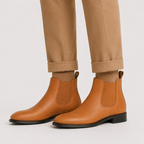 Tan leather Chelsea ankle boots worn with cuffed tan trousers against a light gray studio background.