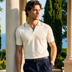 Man wearing a white polo shirt with black trim and dark pants, standing outdoors with columns and greenery in the background.