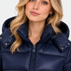 A close-up of a woman wearing a navy blue puffer jacket with a high padded collar and front zipper, against a light gray studio background.
