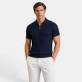 Man wearing a navy ribbed knit polo shirt with white trousers, standing against a light gray background.