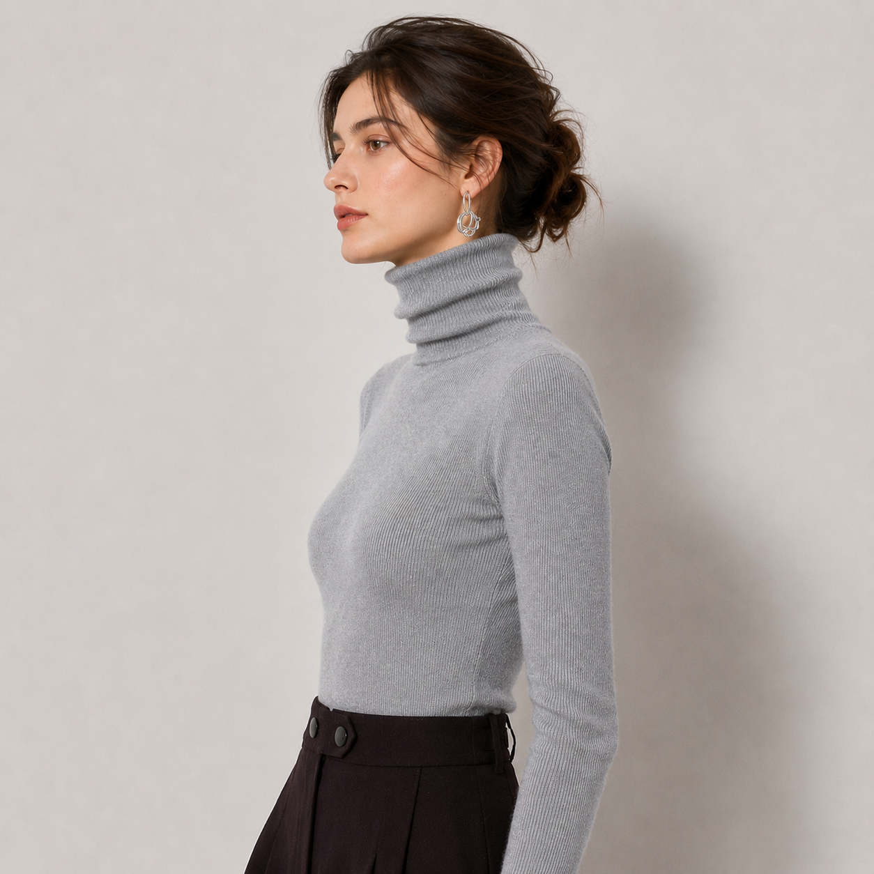 A woman in profile wearing a light gray ribbed turtleneck sweater and dark brown pleated trousers, standing against a neutral light gray wall.