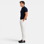 Side view of a man wearing a navy ribbed knit polo shirt with white trousers and navy loafers against a light gray background.