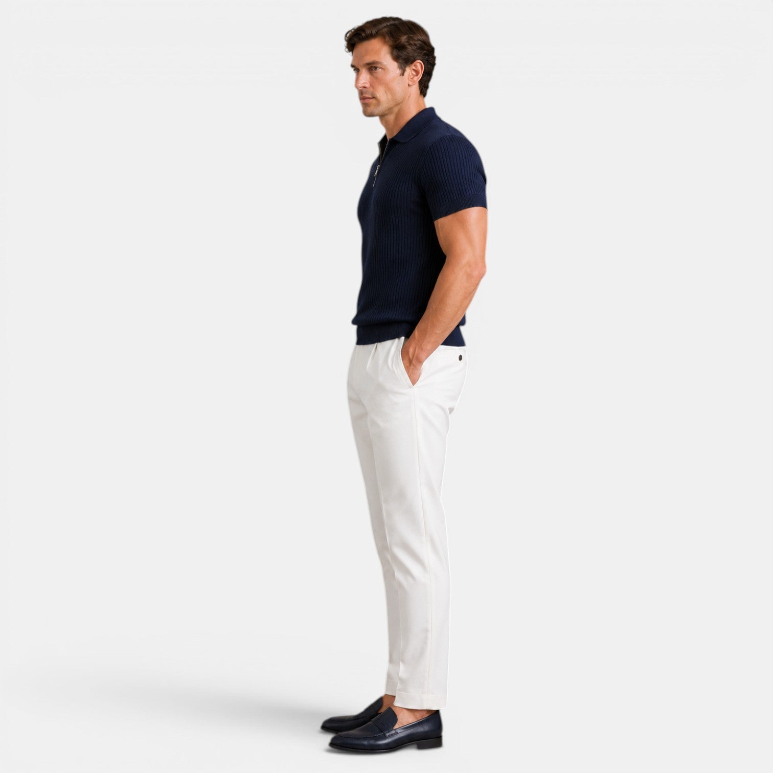 Side view of a man wearing a navy ribbed knit polo shirt with white trousers and navy loafers against a light gray background.