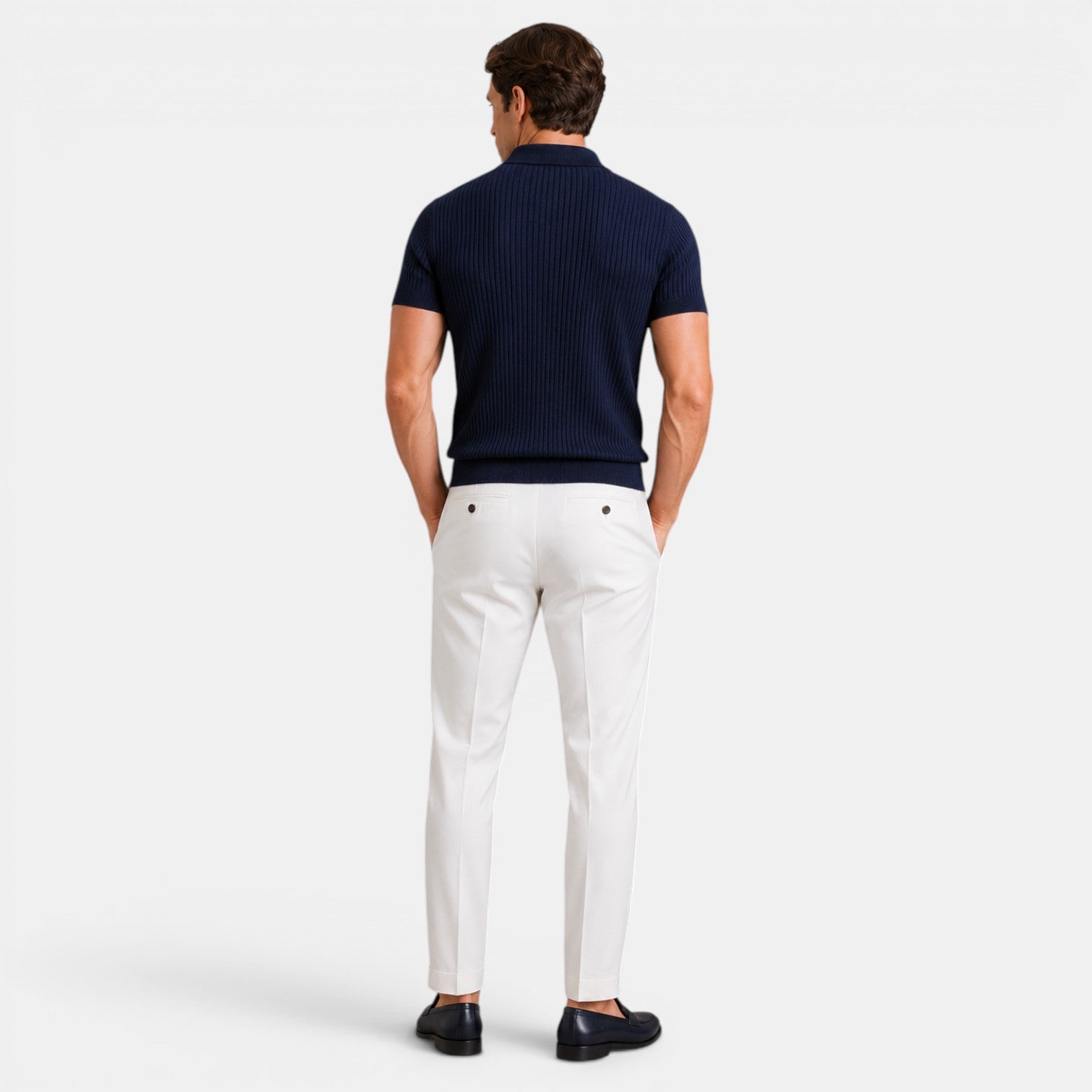 Back view of a man wearing a navy ribbed knit polo shirt with white trousers and navy loafers against a light gray background.