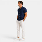 Side view of a man wearing a navy short-sleeve button-up shirt with white drawstring trousers and white sneakers against a light gray background.