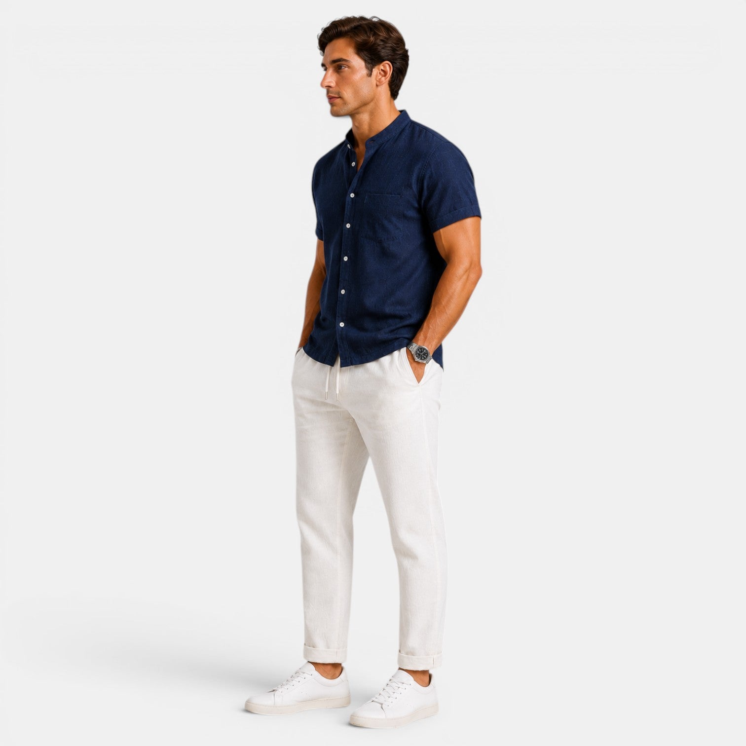 Side view of a man wearing a navy short-sleeve button-up shirt with white drawstring trousers and white sneakers against a light gray background.