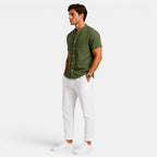 Side view of a man wearing an olive green short-sleeve button-up shirt with white drawstring trousers and white sneakers against a light gray background.