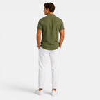 Back view of a man wearing an olive green short-sleeve button-up shirt with white drawstring trousers and white sneakers against a light gray background.