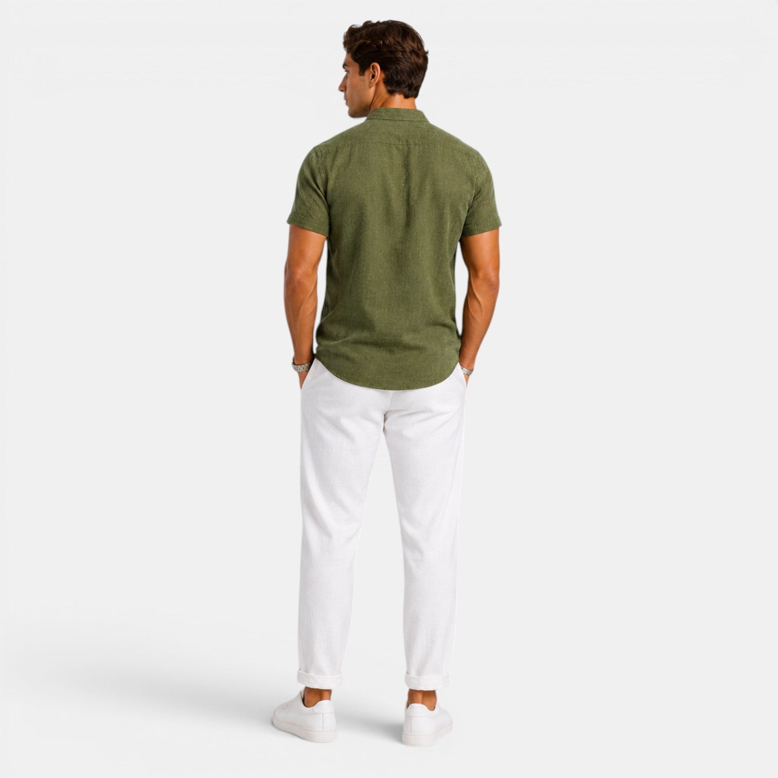 Back view of a man wearing an olive green short-sleeve button-up shirt with white drawstring trousers and white sneakers against a light gray background.