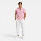 Man wearing a pink short-sleeve button-up shirt with white drawstring trousers and white sneakers, standing against a light gray background.