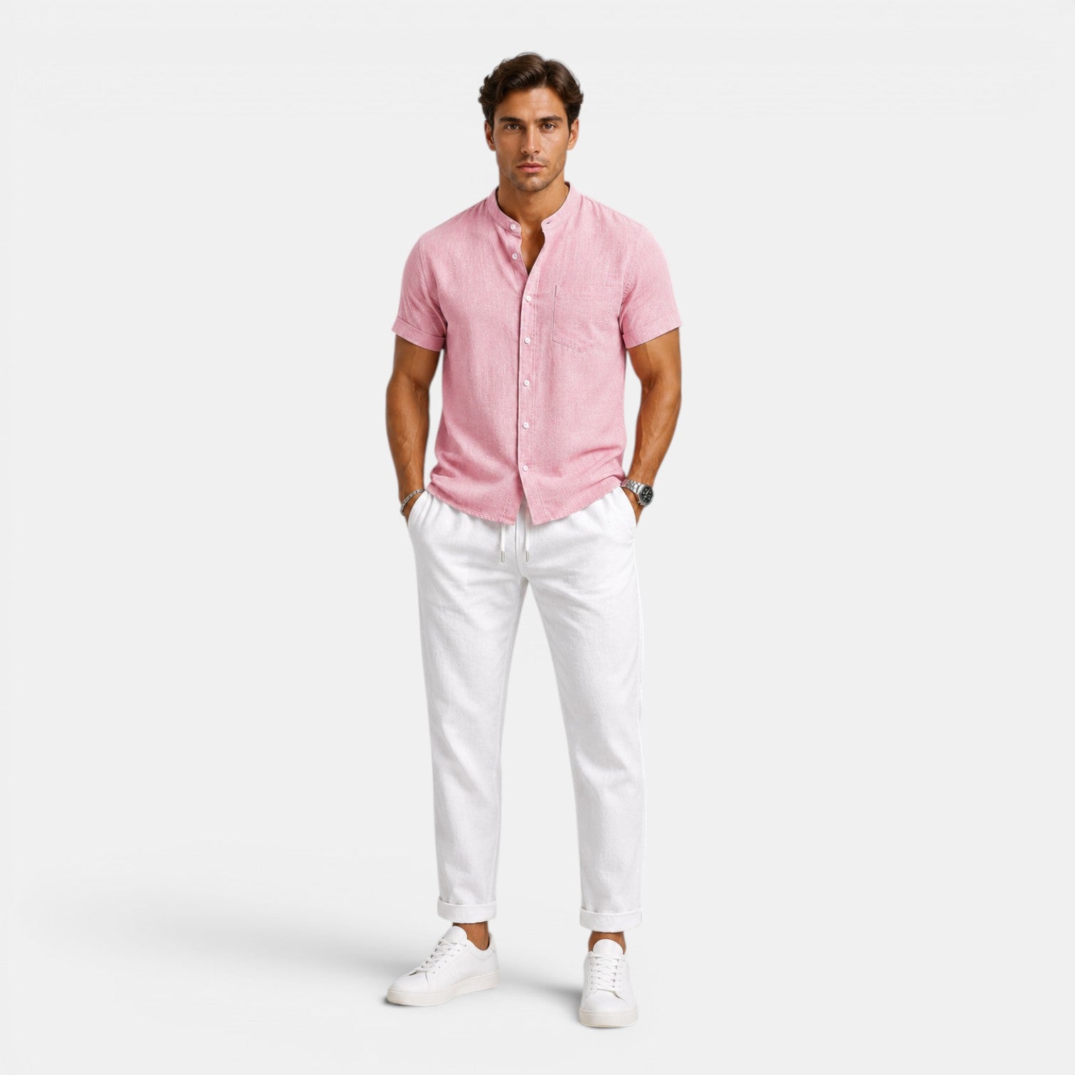 Man wearing a pink short-sleeve button-up shirt with white drawstring trousers and white sneakers, standing against a light gray background.