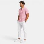 Side view of a man wearing a pink short-sleeve button-up shirt with white drawstring trousers and white sneakers against a light gray background.