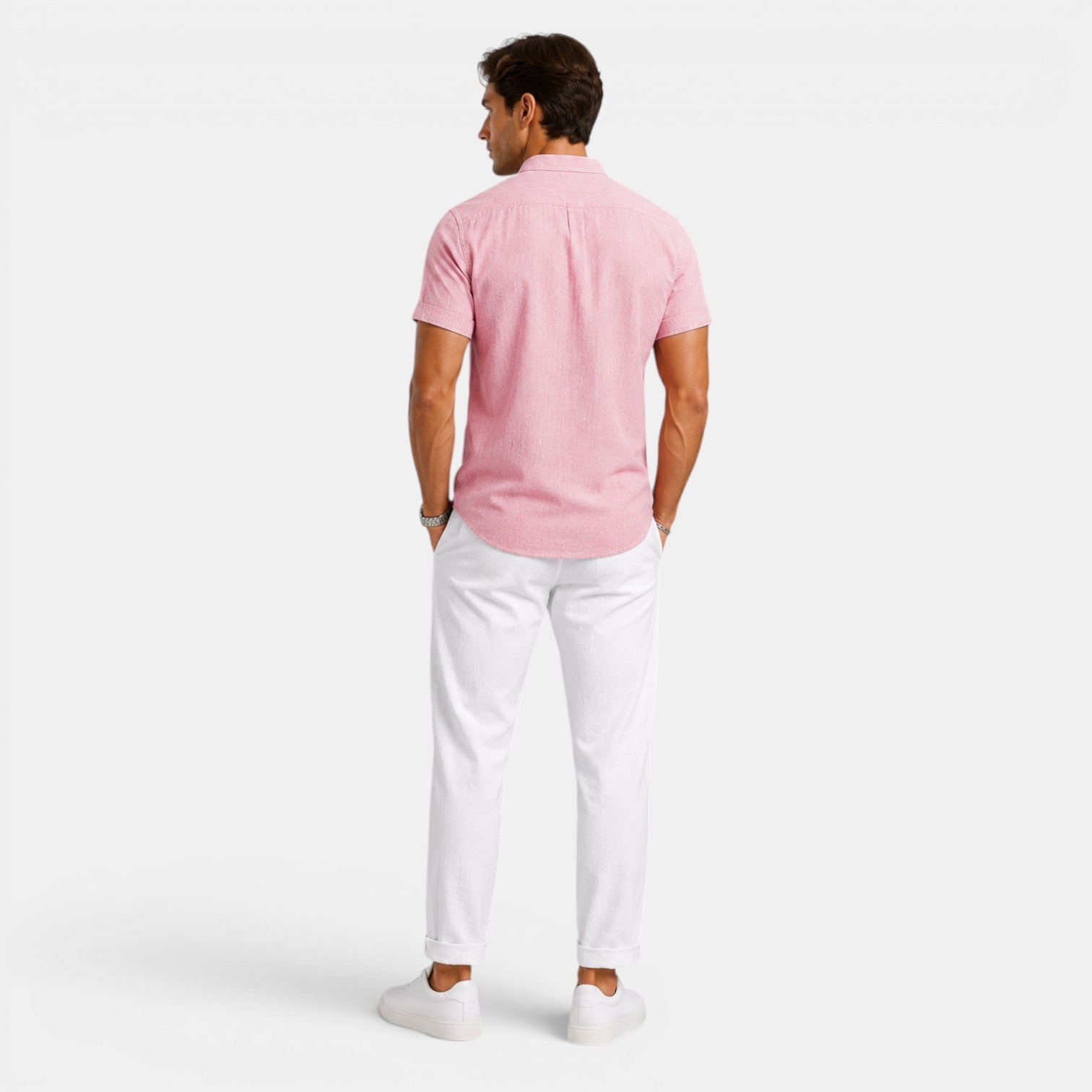 Back view of a man wearing a pink short-sleeve button-up shirt with white drawstring trousers and white sneakers against a light gray background.