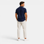 Back view of a man wearing a navy short-sleeve button-up shirt with white drawstring trousers and white sneakers against a light gray background.