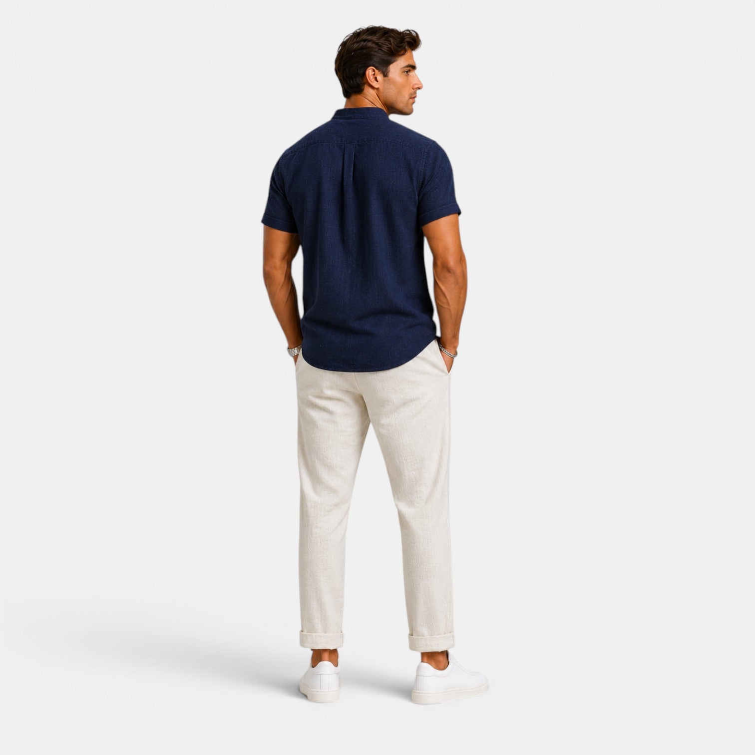 Back view of a man wearing a navy short-sleeve button-up shirt with white drawstring trousers and white sneakers against a light gray background.