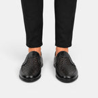 Black perforated leather loafers worn with black trousers, shown from the front.