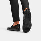 Black perforated leather loafers worn with black trousers, featuring a textured rubber outsole.