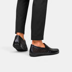 Black perforated leather loafers worn with black trousers, shown from a rear side angle.