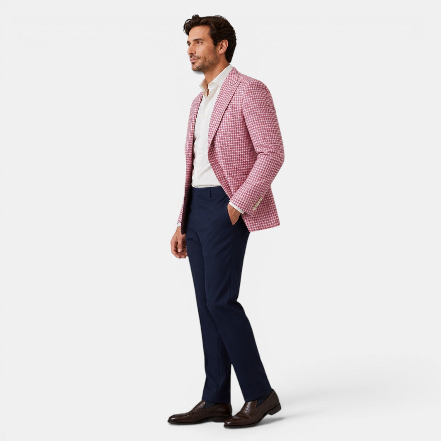 Full-length side view of a young man wearing a pink checked blazer, white dress shirt, navy trousers, and brown loafers.