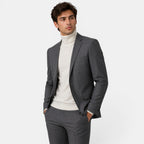 Young man in a charcoal gray suit with a cream turtleneck sweater, posing with hands in pockets.